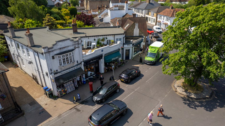 An aerial view of Penge High Street showing a mix of shops with large windows and awnings, some featuring signage. The pavement in front of the stores is clean and lined with a few pedestrians, includ